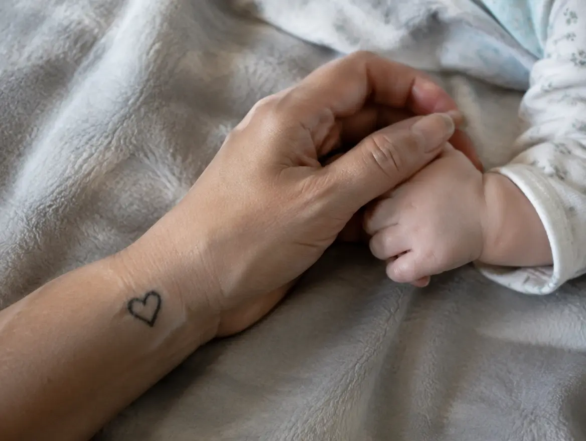 En förälders underarm med en liten hjärttatuering håller en bebis hand